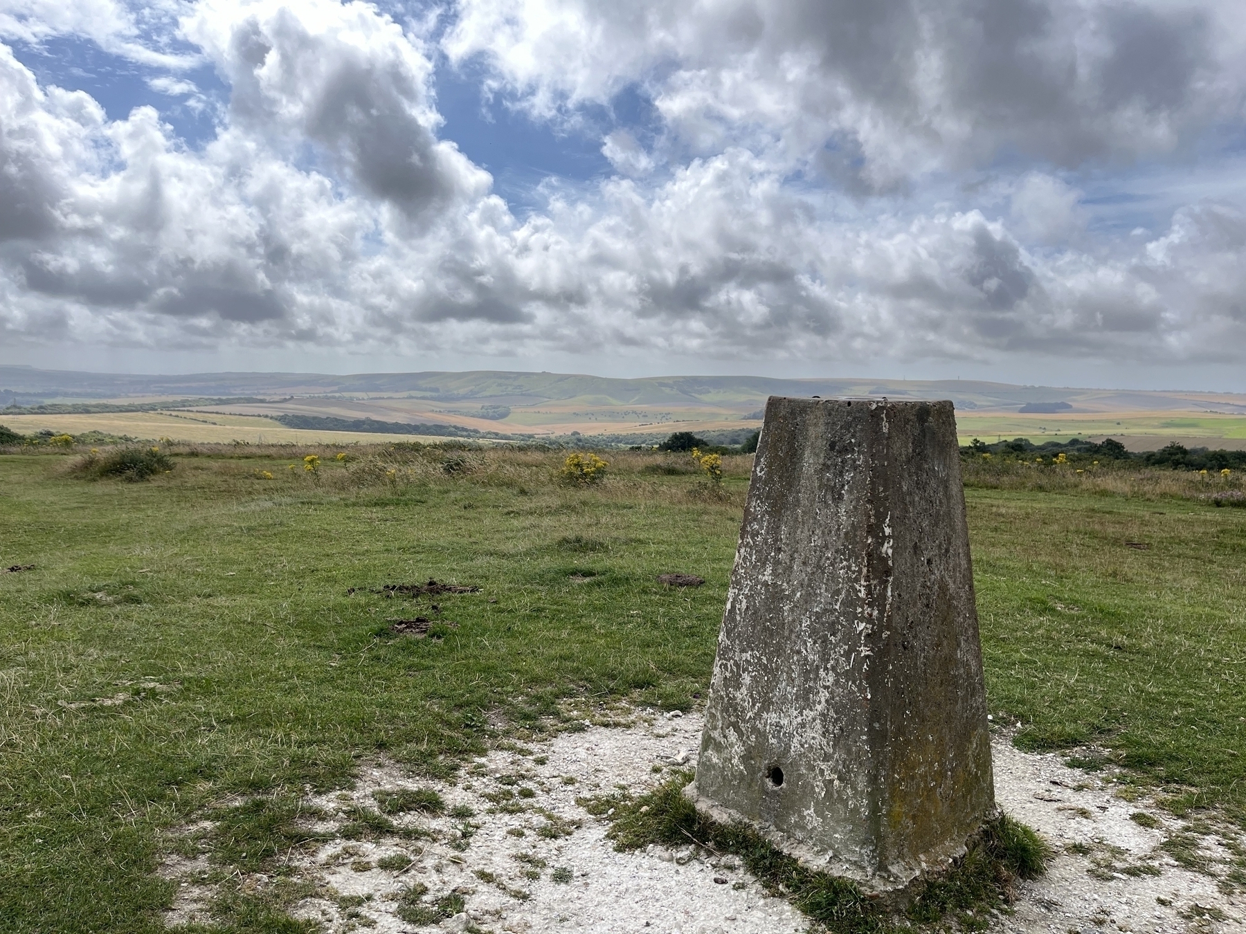 A triangular concrete trig point in the foreground, a view of the South Downs in the distance, and dramatic clouds with glimpses of blue sky overhead. 